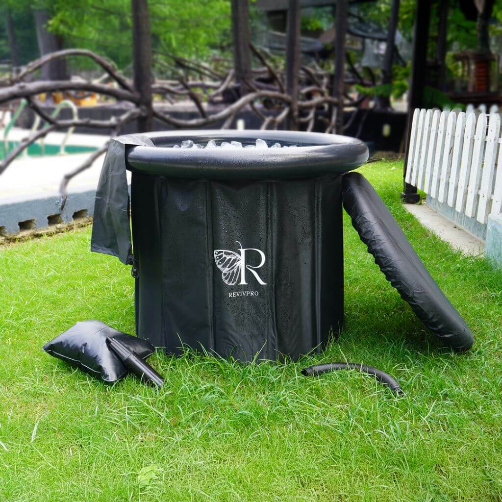 A portable foldable ice bath tub set on a grassy area, primarily black in color with a logo on the side, accompanied by a cover lid, hand pump, and drainage hose.
